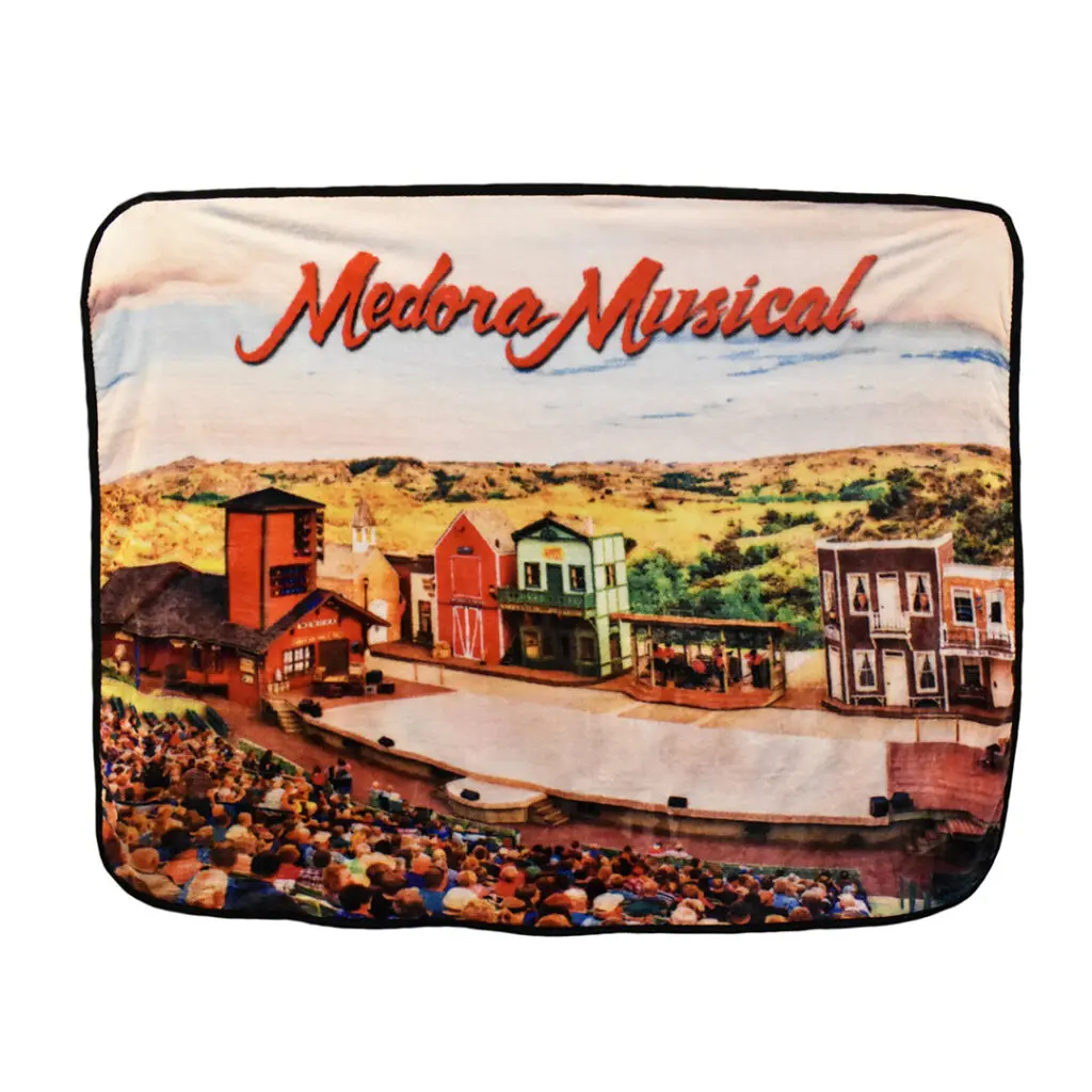 A plush blanket which displays a picture of a full audience sitting in the Burning Hills Amphitheatre overlayed with red text which reads "Medora Musical" which can be found in stores in Medora, North Dakota