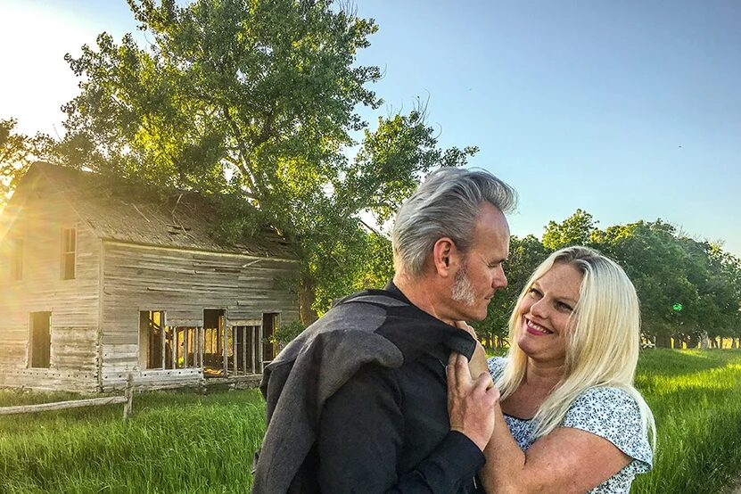 A couple poses in front of an old house, green trees, and a field, a cover image for the American-Gospel Band the Floras, playing in Medora, North Dakota May 11-12, 2024