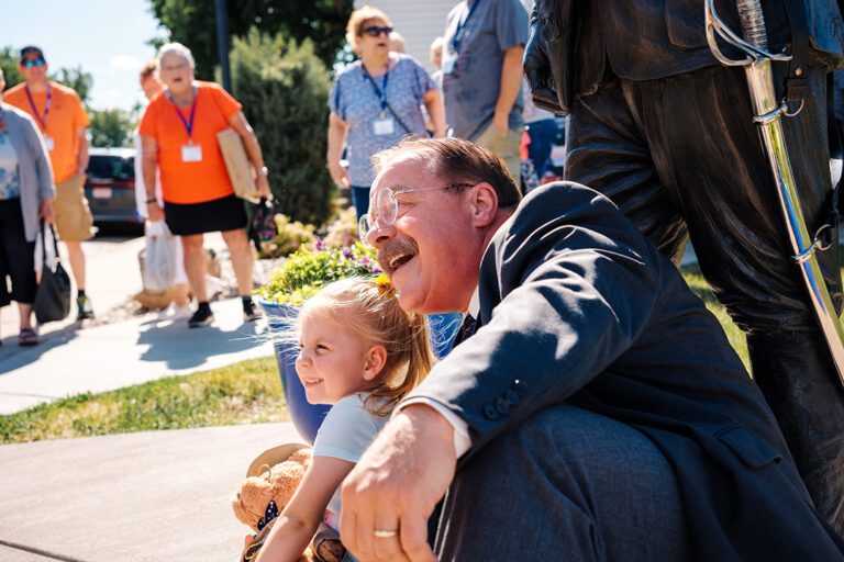 Joe Wiegand dressed as Theodore Roosevelt sits with a young girl holding a Teddy Bear in front of a Theodore Roosevelt statue. Guests gather around the Old Town Hall Theater, a historic theater in downtown Medora, North Dakota