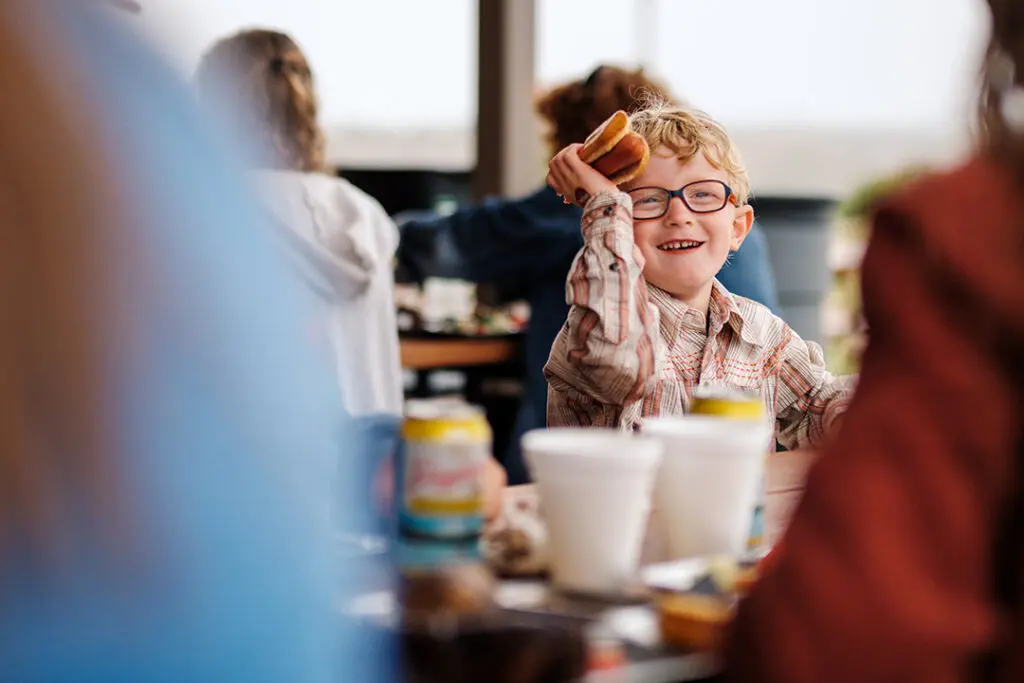 A kids holds up a hotdog and smiles as he sits at a table with his family at the Pitchfork Steak Fondue, a dining attraction in Medora, North Dakota