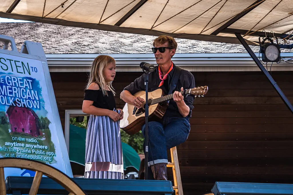 A man sits on a stool with a guitar and sings into a mic stand with a young girl on stage at the Great American Folk Show, a free show in Medora, North Dakota