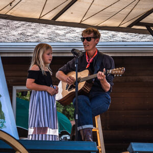 A man sits on a stool with a guitar and sings into a mic stand with a young girl on stage at the Great American Folk Show, a free show in Medora, North Dakota