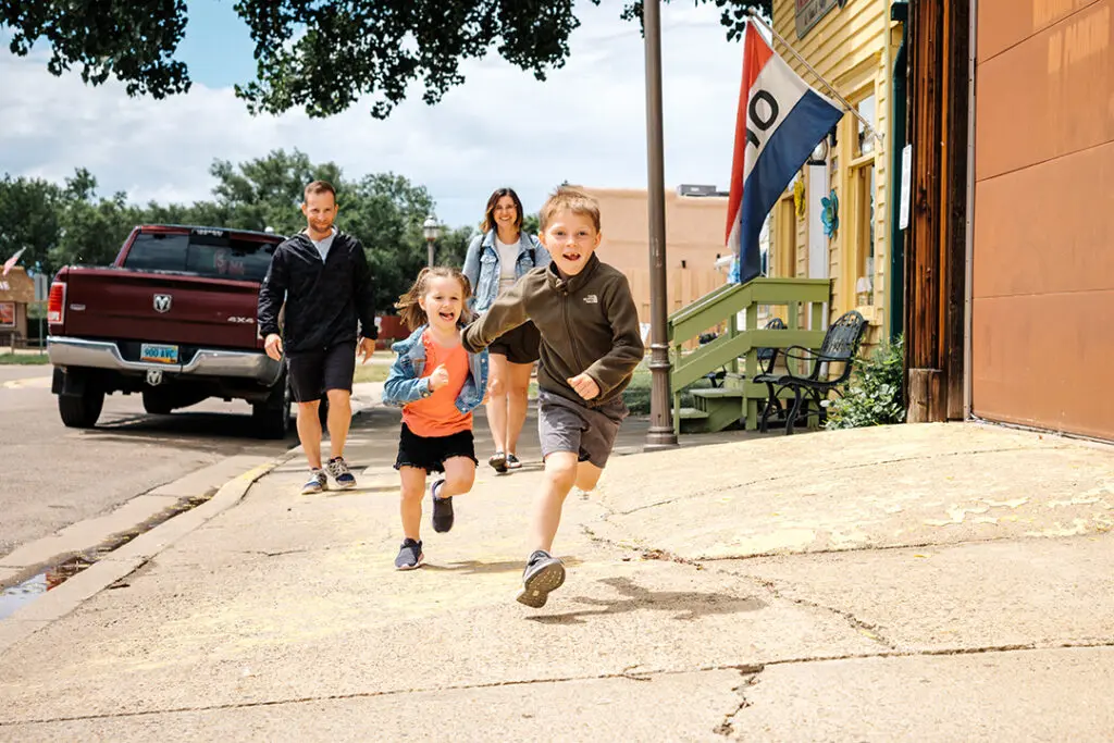 Two smiling kids run down the sidewalk in downtown Medora, North Dakota. Their parents follow, also smiling.