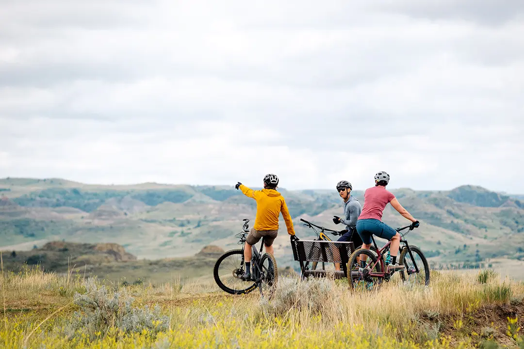 Three bikers rest on the top of a trail on the Maah Daah Hey, a popular outdoor attraction for mountain biking and hiking in Medora, North Dakota. A biker dressed in yellow points out to a distant view of the Badlands.