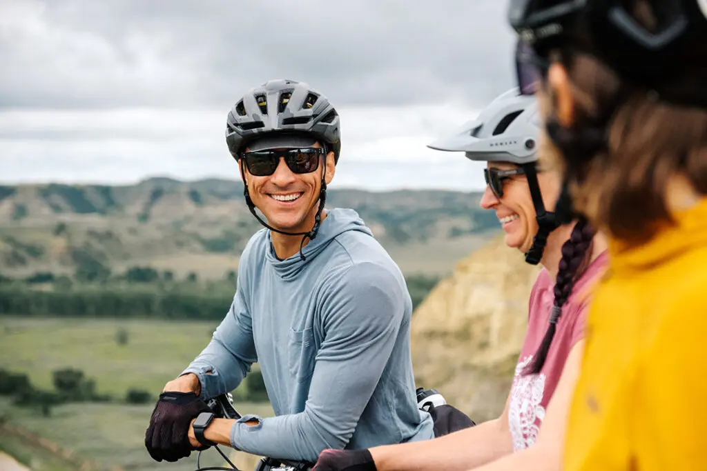 A man on a bike in a bike helmet smiles towards two bikers on the Maah Daah Hey Trail, a popular outdoor attraction for mountain biking and hiking in Medora, North Dakota