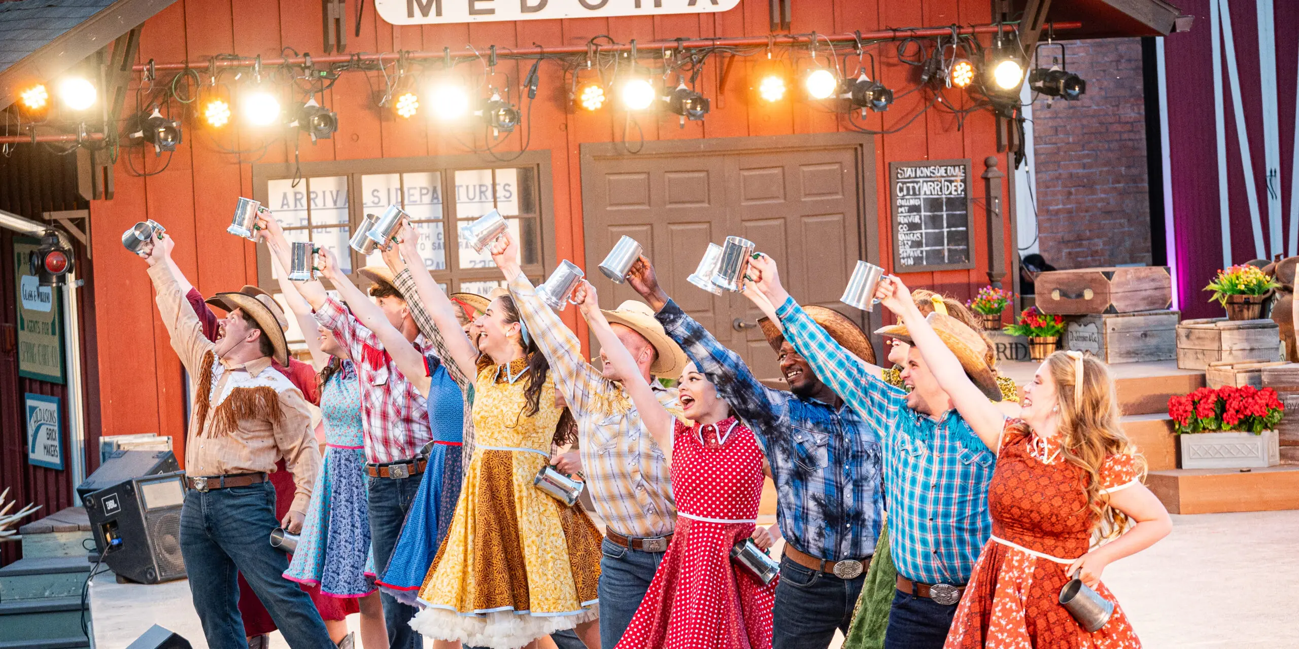 A group of performers in colorful cowboy costumes lift mugs and smile on stage at the Medora Musical, a popular entertainment attraction in Medora, North Dakota