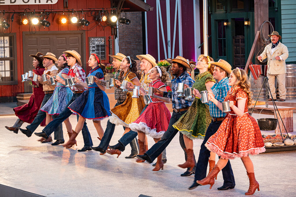 Performers in colorful dresses and cowboy hats dance in a line holding mugs as Theodore Roosevelt watches on stage at the Medora Musical, a popular entertainment attraction in Medora, North Dakota