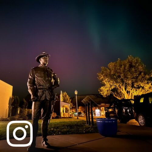 A life size Teddy Roosevelt bronze statue stands against a sky lit by purple and green northern lights at Theodore Roosevelt National Park, Medora, ND